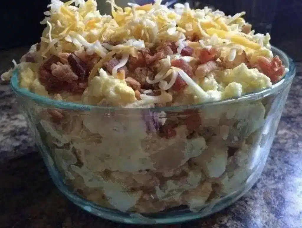 A serving bowl of loaded baked potato salad garnished with extra cheddar, bacon bits, and chives