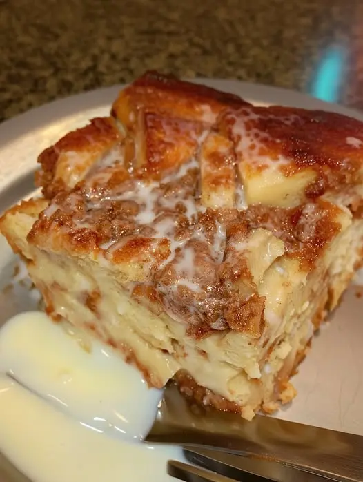 A serving of cinnamon roll bread pudding on a white plate, topped with a drizzle of white icing.