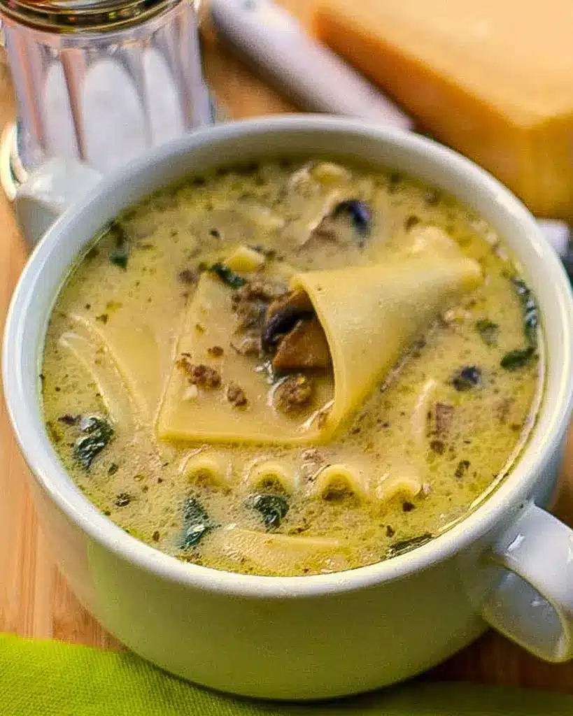 A pot of creamy alfredo lasagna soup being stirred with a wooden spoon, showing noodles, chicken, and spinach.
