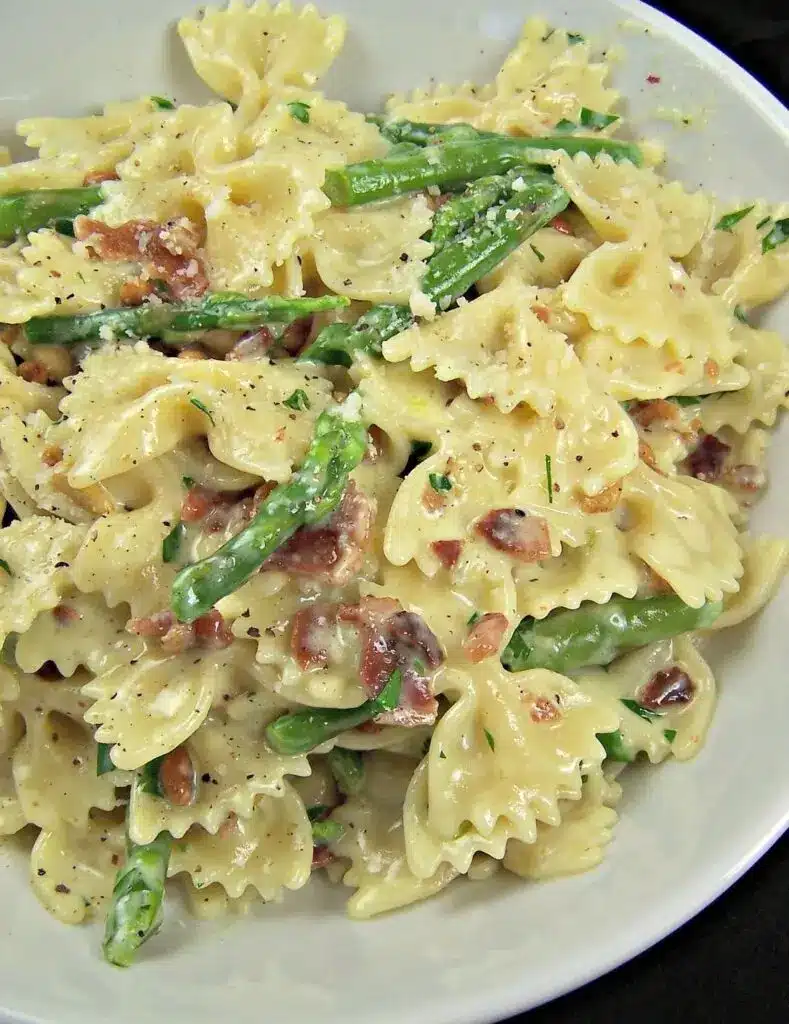 A beautiful plate of creamy pasta with green asparagus, bacon bits, and a garnish of fresh herbs and lemon.