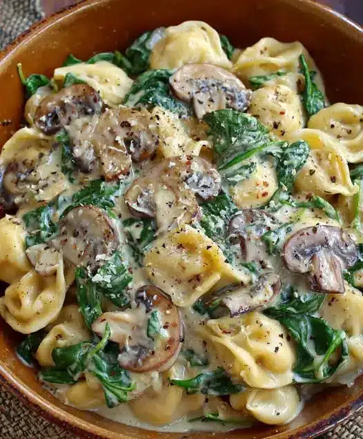 A close-up shot of creamy tortellini with wilted spinach and browned mushrooms in a white bowl