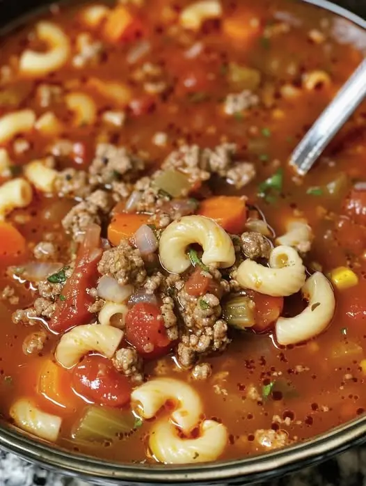 A bowl of hot beef macaroni soup garnished with fresh parsley next to a piece of crusty bread