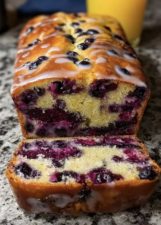 A whole loaf of blueberry bread sitting on a wooden cutting board with a slice removed.