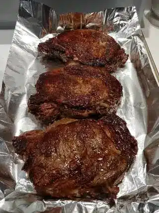 A sliced medium-rare ribeye steak on a platter, garnished with roasted garlic and herbs.